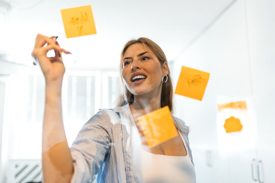 Creative businesswoman writing on glass wall with sticky notes. Smiling young female employee brainstorming project ideas using adhesive memos in modern office. Corporate agile workspace concept.