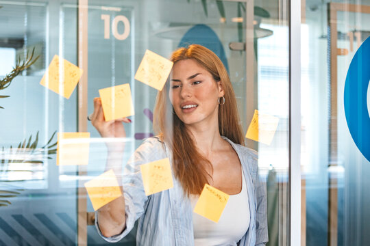 Happy businesswoman writing notes on glass wall in office. Smiling young female entrepreneur planning project with sticky memos during agile meeting in modern workspace. Corporate lifestyle concept.