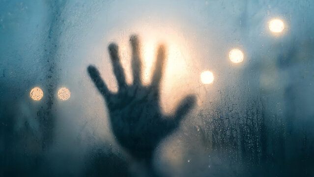 A dark hand silhouette presses against a fogged glass surface, with warm blurred bokeh lights in the background, creating a mysterious moody scene.