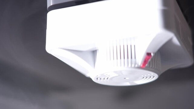 Close-up of ceiling-mounted fire alarm. White casing with red indicator light. Visible vents on the device. Suspended from ceiling structure. Typical building safety equipment
