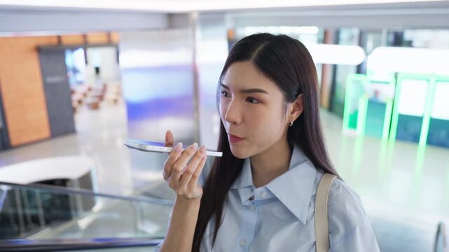 Young Asian woman speaking a voice message into her smartphone while riding an escalator in a modern building, staying connected on the move during her commute.