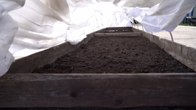 Wooden garden bed holds rich dark soil. White fabric covers the bed. Fabric shields soil from rain and debris. Ideal for spring planting preparation. Shows garden readiness