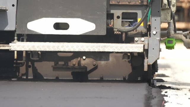 Close-up of road roller's metal plow gliding over fresh asphalt. Green sensor monitors pavement texture and quality. Hot mix spreads smoothly on the road surface