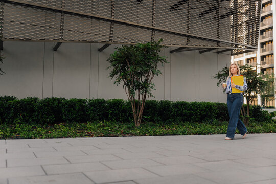 Blonde woman walking with coffee and folder in urban area