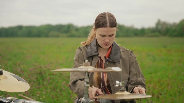 Young Caucasian female drummer assembling drum kit in open green field, adjusting cymbals in slow motion with zoom out. Suitable for indie music marketing and youth lifestyle campaigns