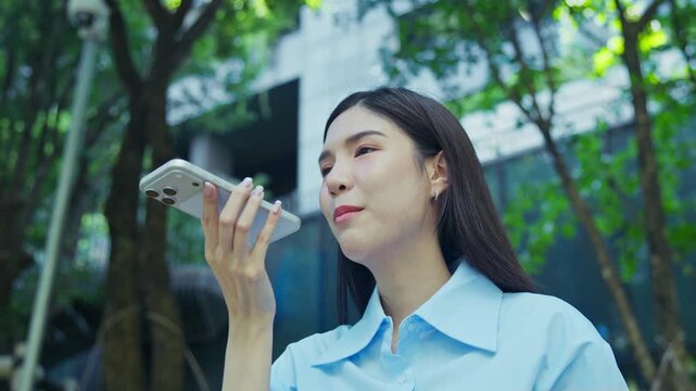 Young Asian woman speaks into her smartphone to record a voice message while standing outside among green trees, gesturing with her free hand as she talks.