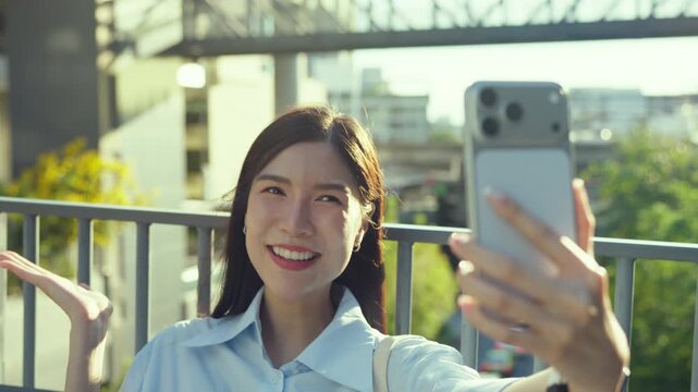 Asian businesswoman holds up her smartphone for a video call at the railing of an elevated walkway, backlit by warm afternoon sun.