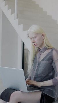 Stylish albino lady working with laptop computer typing sitting on stairs in elegant luxury apartment. Modern technology and house architecture concept.