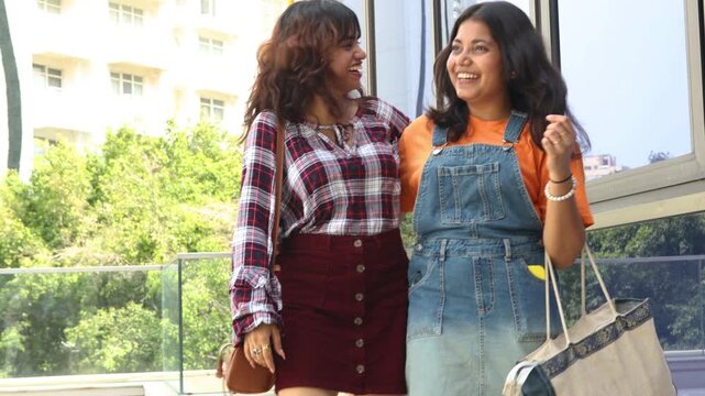 Two beautiful young indian Gen Z female friends walking together holding shopping bags in hand laughing and pointing at something with expressions of surprise and joy. modern city lifestyle in india