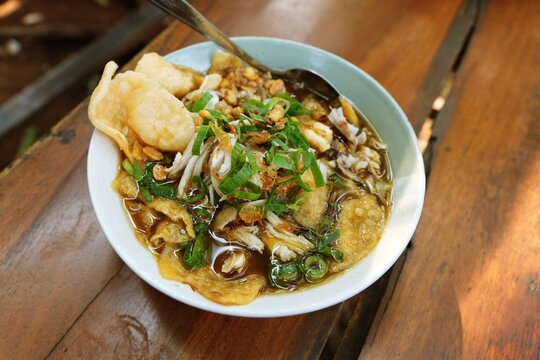 Traditional Indonesian bubur sop served in a white bowl with vegetables, savory toppings and crispy crackers, photographed on a yellow table in a casual food setting.