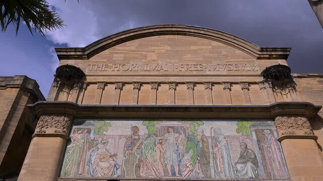 Ornate stone entrance of the Horniman Museum and Gardens with a colorful mosaic frieze framed against a dramatic cloudy sky.
