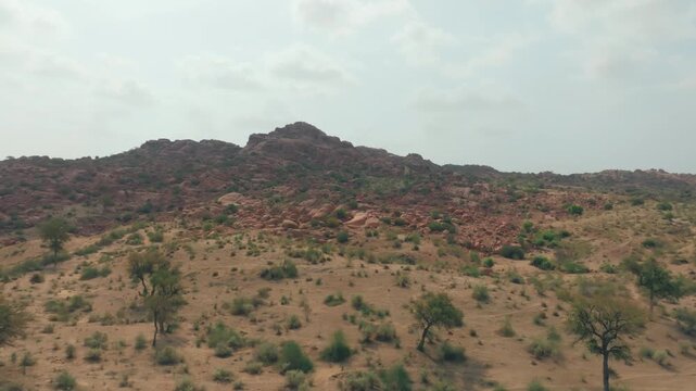 Drone view of a rugged rocky hill rising from the Thar Desert in Sindh, Pakistan, with scattered thorn trees and sparse scrub across the dry sandy slopes.