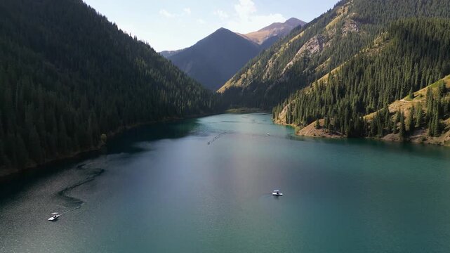 A drone flies over the beautiful Kolsai Lake, surrounded by pine forest and the Tien Shan Mountains. It's a popular tourist attraction in Kazakhstan