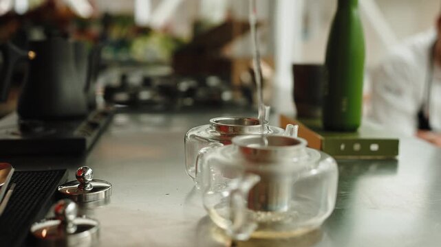 Pouring steaming hot water from a gooseneck kettle into a glass teapot with infuser, brewing fresh herbal tea at a kitchen counter