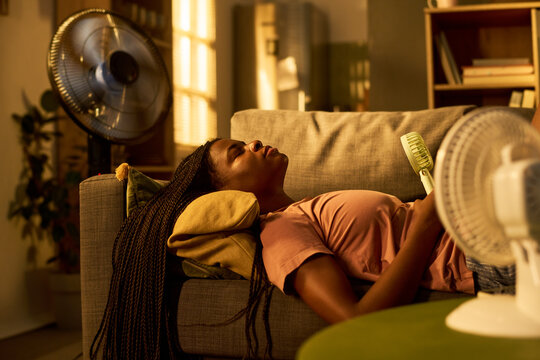 Young African American woman lying on sofa cooling off with electric fans during summer heatwave. Image suitable for climate change awareness and cooling appliance marketing