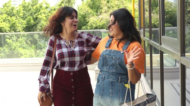 Two beautiful young indian Gen Z female friends walking together holding shopping bags in hand laughing and pointing at something with expressions of surprise and joy. modern city lifestyle in india