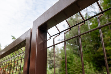 Fototapeta premium Low angle view of a brown metal 3D mesh wire fence gate section on a blurred forest and cloudy sky background