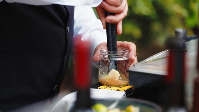 Bartender's hands mashing fresh fruit inside a small glass jar with a muddler, preparing a craft cocktail