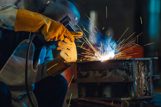 Expert metalworker in full safety gear performing heavy arc welding on a steel construction part, creating smoke and glowing flying sparks inside an industrial manufacturing workspace.