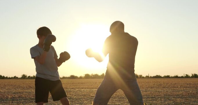 Father and son practice boxing in open farm field at sunset. Young athlete learns punches from parent during outdoor training. Warm backlight captures family fitness, discipline, and teamwork.