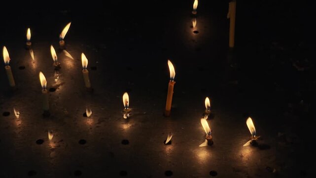 multiple thin, yellow wax candles burning in a dark setting, likely inside a church or place of prayer