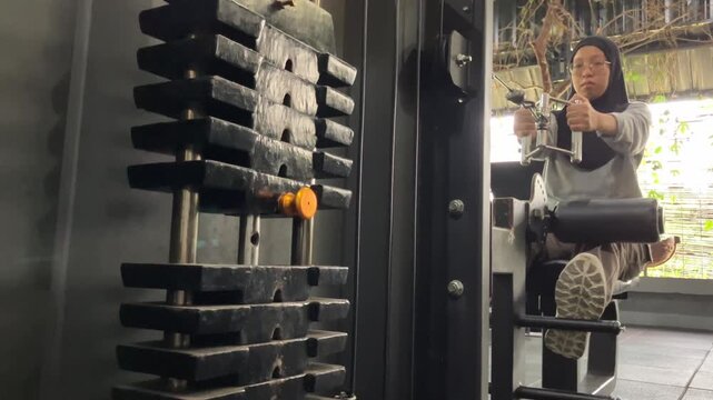 Slow motion video of a hijab-wearing woman performing a strength training exercise on a gym cable machine, focusing on the moving weight stack.