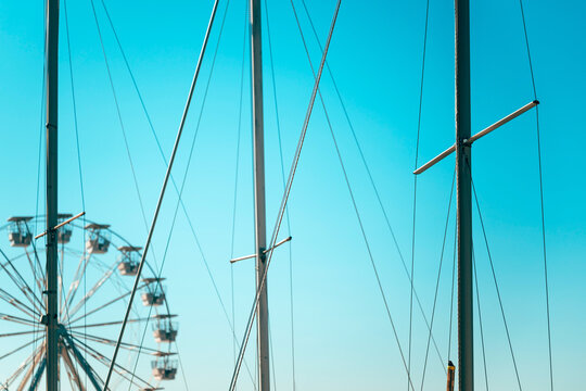 Sailboat masts and rigging in a coastal harbor with a blurred ferris wheel against a bright turquoise sky.