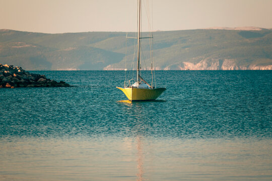 Anchored yellow sailboat on tranquil sea with coastal mountains in the background. Concept of Mediterranean summer travel, freedom, and sailing.