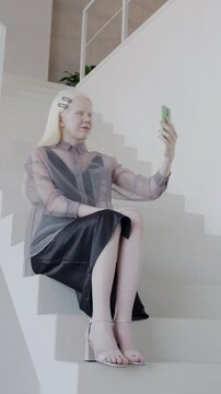 Attractive albino woman making online video call sitting on stairs in luxury house hall. Distance communication and stylish interior concept.
