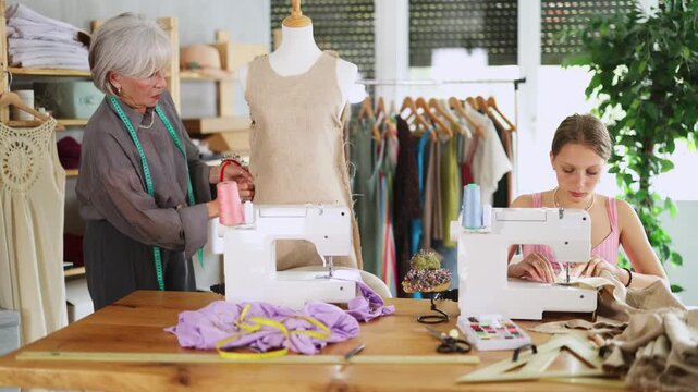Skillful young tailoress using sewing-machine while mature woman working with mannequin in sewing workshop . High quality 4k footage