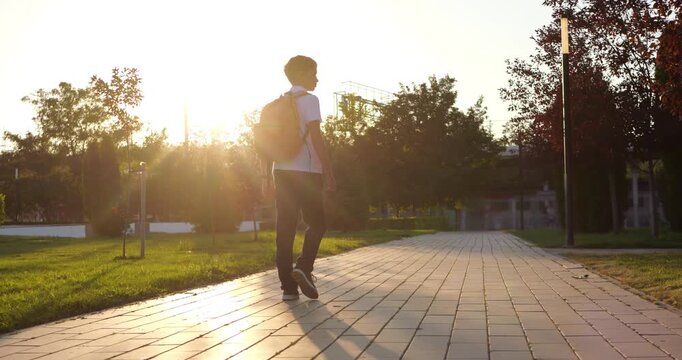 Schoolboy with backpack walks along paved park path at sunset. Student enjoys peaceful outdoor journey through urban greenery, representing education, independence, childhood and growing up.