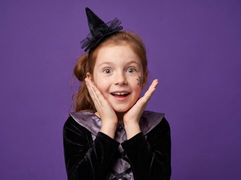 Happy little girl dressed as a witch for Halloween, looking surprised and excited against a purple background.