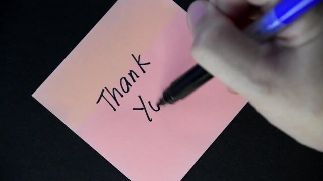 Close up of a female hand writing a thank you note on a pink sticky note.