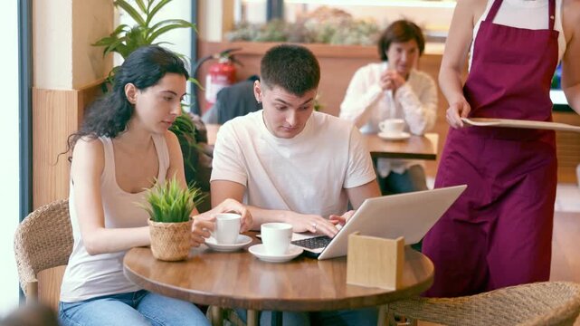 Young man and a woman are sitting at a coffee shop table and discussing the details of a project while looking at a laptop. Man with a woman at a business meeting over a cup of hot coffee. . High