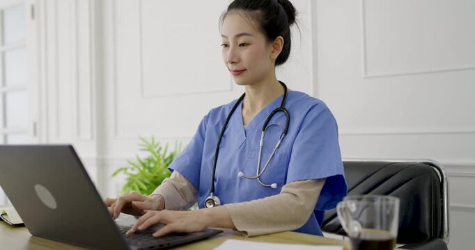 Portrait of female woman doctor healthcare professional in medical scrubs typing on a laptop, representing digital healthcare, patient records, and tele medicine services.