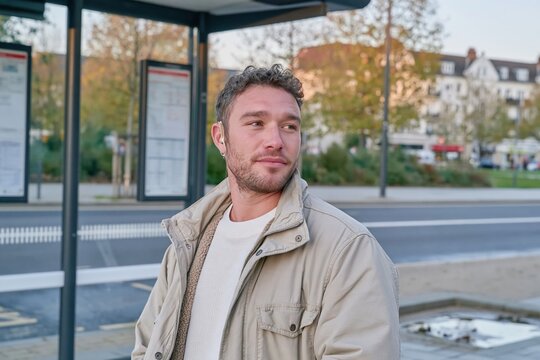 Man waiting at a bus stop shelter on a dim evening, looking toward the road for transit and travel campaigns