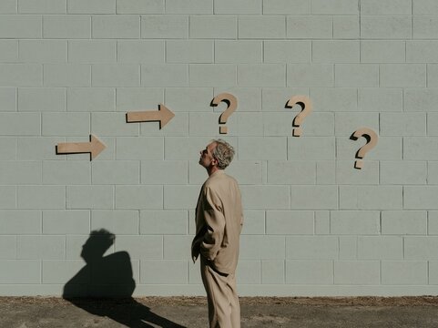 Midterm elections concept featuring a pensive man looking at wooden arrows and question marks on a pale grey cinder-block wall, copy space