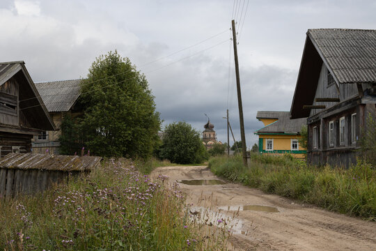 A summer rural landscape. Wooden houses line the village street. An Orthodox church with a leaning cross on its dome is visible in the distance. Pozharishche village, Vologda Oblast, Russia.