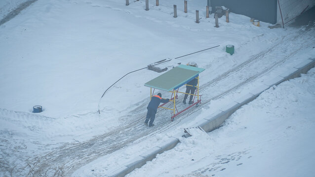 During the harsh winter months, a team of diligent workers busily constructs a sturdy shelter amidst a picturesque snowy landscape. They are situated near a building in an area characterized 