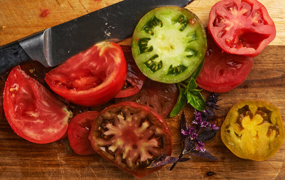 Sliced heirloom tomatoes of different colors and varieties on a wooden cutting board with a knife and purple basil, messy top view food photography