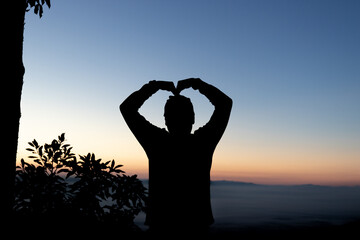 Silhouette of person forming heart gesture with hands against morning sunrise sky. © wannasak