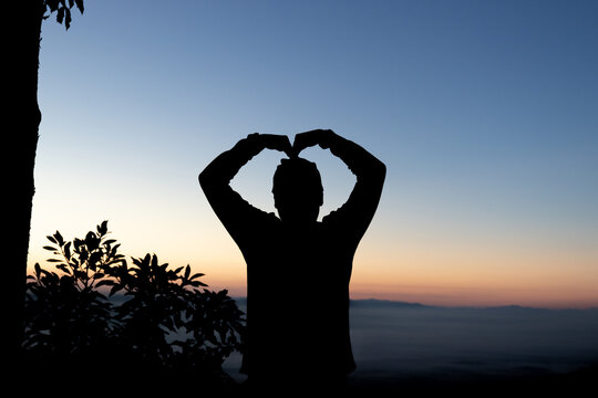 Silhouette of person forming heart gesture with hands against morning sunrise sky.