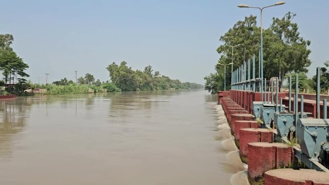 A daytime panoramic video shot of a flowing river next to a water barrage control structure with sluice gates, surrounded by lush green trees and buildings. This stock footage is ideal for themes rela