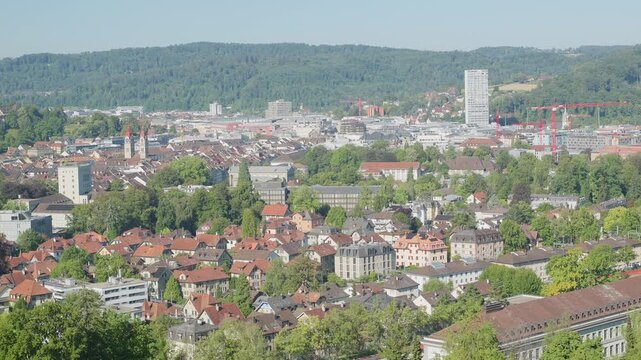A sprawling cityscape nestled among lush green trees. A mix of traditional buildings with red-tiled roofs and modern structures, including a tall white high-rise, are visible.