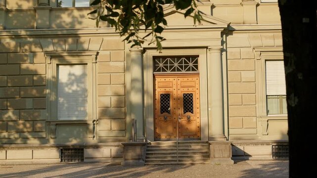 The image shows the entrance to a building with double wooden doors and stone steps. Shadows of leaves and tree branches fall across the facade.