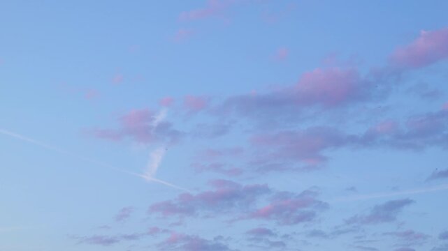 A soft blue sky is filled with scattered clouds tinged with pink and some white contrails. A few tiny birds are visible against the vast expanse.