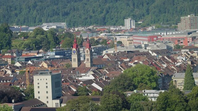 An aerial view captures a densely populated city with red-roofed buildings, a prominent clock tower, and a lush green forest backdrop.