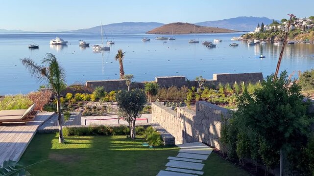 A stunning view of Bitez Bay in Turkey from a luxury resort terrace, with green lawns, stone walls, palm trees, and yachts moored on calm blue waters.