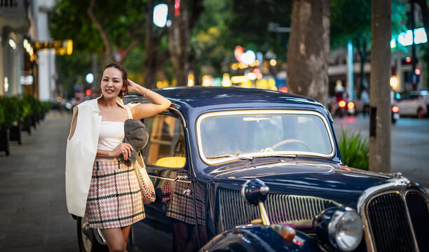 Stylish Asian woman smiles beside a classic car on a city street