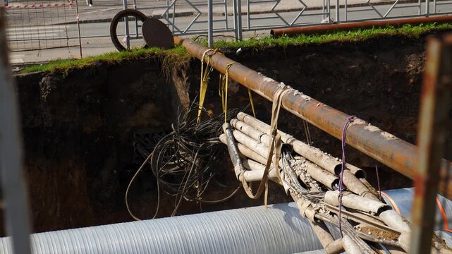 View of construction trench with corrugated pipe and bundle of cables tied to rusty metal pipe. Underground utility repair, urban infrastructure work, mesh fence in foreground 4k video